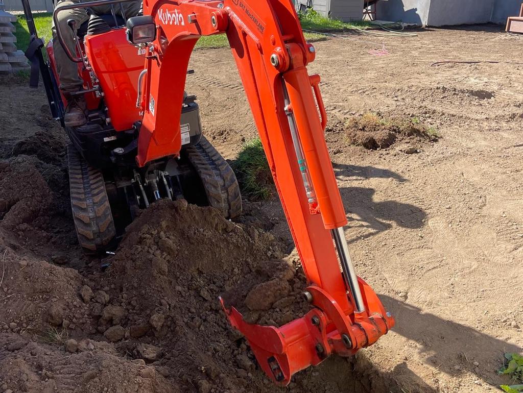 Kubota mini excavator is actively engaged in a precise landscaping task, its arm extended into the soil of a residential garden area, showcasing the early stages of a meticulous outdoor renovation.